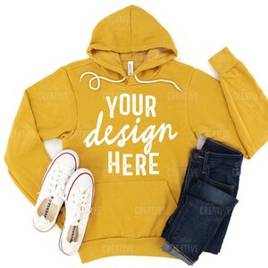 May include: Mustard yellow hoodie with white text that reads "YOUR design HERE". The hoodie is paired with dark blue jeans and white canvas sneakers with red accents. The items are arranged on a white background.