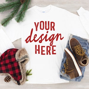 May include: White long-sleeved t-shirt with the text "YOUR design HERE" in red. The shirt is lying flat on a wooden surface with a plaid hat, jeans, and brown shoes.