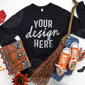 以下が含まれることがあります： 白い文字で「YOUR design HERE」と書かれた黒いスウェットシャツ、ほうき、本、オレンジ色のスニーカー、そして秋の装飾が白い木製の背景に置かれています。