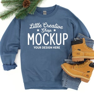 May include: A blue crewneck sweatshirt with the text "Little Creative Shop MOCKUP YOUR DESIGN HERE" is displayed with a pair of tan work boots and blue jeans. A sprig of evergreen and a pinecone are in the upper left corner.