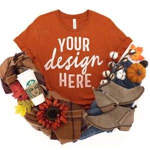 May include: A burnt orange t-shirt with the text "YOUR design HERE" in white. The shirt is styled with a plaid scarf, a coffee cup, denim jeans, and fall-themed decor.