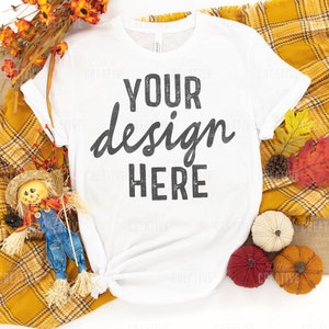 May include: White t-shirt with the text "YOUR design HERE" in black. The shirt is styled with fall-themed decorations, including a yellow plaid blanket, pumpkins, and fall leaves. A scarecrow doll is also included.