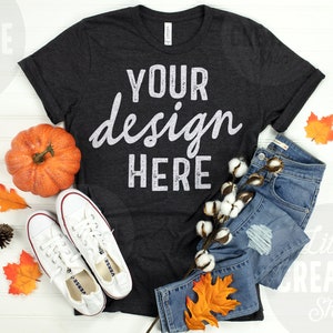 May include: A dark gray t-shirt with the text "YOUR design HERE" in white. The shirt is laid flat on a white wooden surface with a pair of white sneakers, a pumpkin, cotton bolls, blue jeans, and fall leaves.