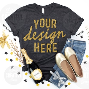 May include: A dark gray t-shirt with gold glitter text that reads "Your design here". The shirt is laying flat on a white surface with gold and black confetti, a bottle of champagne, a pair of gold flats, and blue jeans.