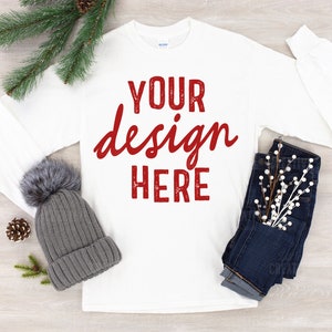 May include: White long-sleeve t-shirt with the text "YOUR design HERE" in red on the front. The shirt is laying flat on a wood surface with a gray beanie, a sprig of greenery, and a pair of blue jeans with white sprigs.