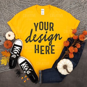 May include: A yellow t-shirt with the text "YOUR design HERE" in black. The shirt is laid flat on a grey surface with a pair of black trainers, a sprig of orange pumpkins, and a white pumpkin.