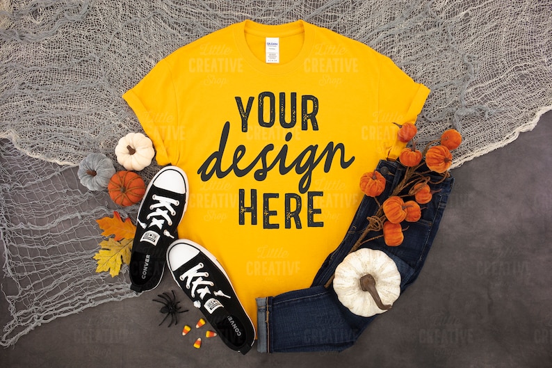 May include: A yellow t-shirt with the text "YOUR design HERE" in black. The shirt is laid flat on a grey surface with a pair of black trainers, a sprig of orange pumpkins, and a white pumpkin.