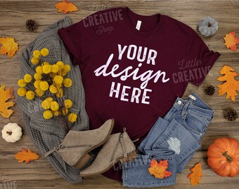 May include: A maroon t-shirt with the text "YOUR design HERE" in white. The shirt is laying on a wooden surface with fall-themed decorations, including a knitted sweater, jeans, boots, and pumpkins.
