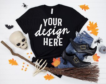 May include: A black t-shirt with the text "YOUR design HERE", alongside blue jeans, black witch boots, and Halloween decorations including a skull, broom, and spiders. Orange autumn leaves and candy corn complete the scene.