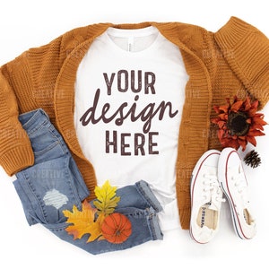 May include: A white t-shirt with the text "YOUR design HERE" in brown lettering. The t-shirt is laid out on a white background with a brown cardigan, blue jeans, fall leaves, a small orange pumpkin, and white sneakers.