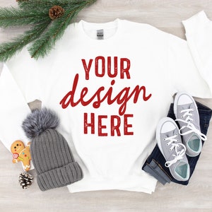 May include: White long-sleeve sweatshirt with the text "YOUR design HERE" in red on the front. The sweatshirt is laid flat on a woodgrain surface with a gray beanie, a gingerbread cookie, a pine branch, and a pair of gray sneakers.
