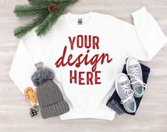 May include: White long-sleeve sweatshirt with the text "YOUR design HERE" in red on the front. The sweatshirt is laid flat on a woodgrain surface with a gray beanie, a gingerbread cookie, a pine branch, and a pair of gray sneakers.