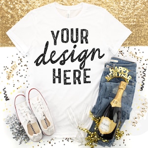 May include: White t-shirt with the text "YOUR design HERE" in black. The shirt is laying flat on a white surface with gold glitter, confetti, a pair of white sneakers, a bottle of champagne, a pair of blue jeans, and a champagne flute.