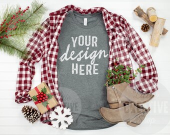 May include: A heather gray t-shirt with the text "YOUR design HERE" in white. The shirt is laid flat on a white surface with a red and white plaid shirt, a small gift box, a pine cone, a snowflake, and a pair of brown boots.