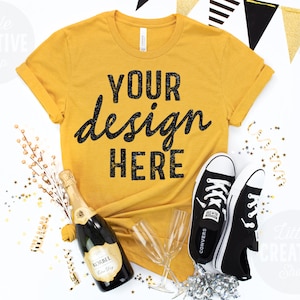 May include: A mustard yellow t-shirt with the text "YOUR design HERE" in black glitter lettering. The shirt is laid flat on a white surface with a bottle of champagne, two champagne flutes, and a pair of black sneakers.