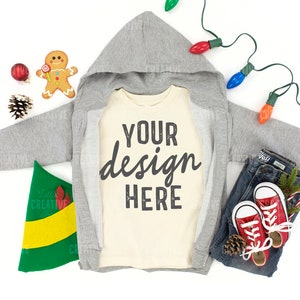 May include: A gray hooded sweatshirt with a cream colored t-shirt underneath. The t-shirt has the text "YOUR design HERE" printed on it. The image is set against a white background with Christmas decorations, including a gingerbread man, a pine cone, a red ornament, and a string of Christmas lights.