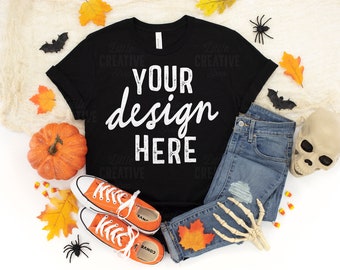 May include: A black t-shirt with the text "YOUR design HERE", alongside blue jeans, orange trainers, and Halloween decorations including a pumpkin, leaves, and a skull. A seasonal apparel flat lay.