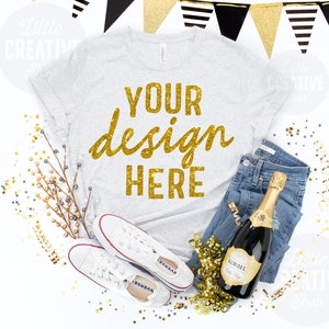 May include: A gray t-shirt with the text "YOUR design HERE" in gold glitter lettering. The shirt is laid flat on a white background with a bottle of champagne, jeans, and white sneakers. There are gold and silver confetti scattered around the shirt.