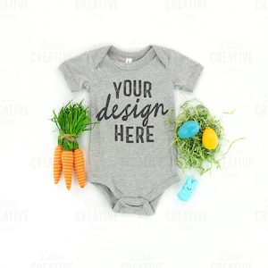 May include: A grey baby onesie with the text "YOUR design HERE" in black. The onesie is surrounded by Easter-themed decorations, including carrots, a nest of grass with blue and yellow eggs, and a blue bunny.