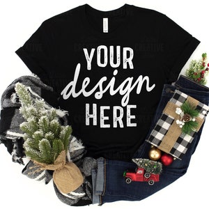 May include: Black t-shirt with the text "YOUR design HERE" in white. The shirt is surrounded by a plaid blanket, a small Christmas tree, a pair of blue jeans, a gift box, and Christmas ornaments.