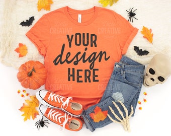 May include: Orange t-shirt with the text "YOUR design HERE" in black. The shirt is surrounded by autumn-themed items, including a pumpkin, leaves, and a skeleton hand.