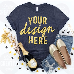 May include: Navy blue t-shirt with gold glitter text that reads "YOUR design HERE". The shirt is styled with a pair of blue jeans, gold flats, and a champagne bottle, with gold and black star confetti.