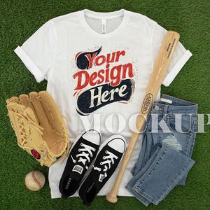 May include: A white t-shirt with the text "Your Design Here", alongside a baseball bat, jeans, a baseball glove, a baseball, and black trainers. The items are arranged on a green surface.