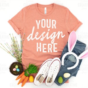 May include: A coral colored t-shirt with the text "YOUR design HERE" in white. The t-shirt is laid flat on a white surface with a pair of white sneakers, a pair of blue jeans, a bunny ear headband, a nest with colorful Easter eggs, and green grass.