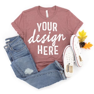 May include: A mauve t-shirt with the text "YOUR design HERE" in white. The t-shirt is laid flat on a white background with a pair of blue jeans and white trainers.