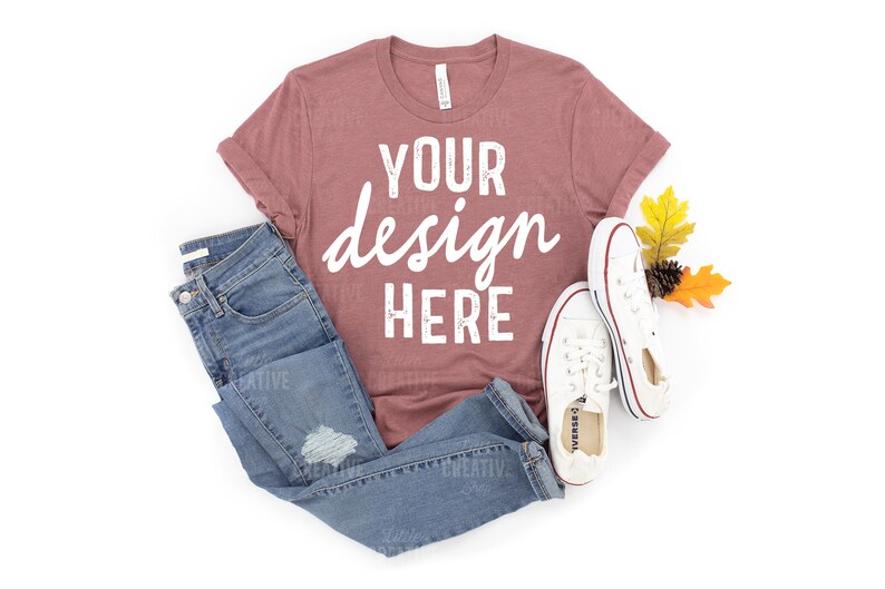 May include: A mauve t-shirt with the text "YOUR design HERE" in white. The t-shirt is laid flat on a white background with a pair of blue jeans and white trainers.