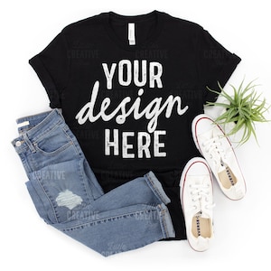 May include: Black t-shirt with the text "YOUR design HERE" in white. The shirt is laid flat on a white background with a pair of blue jeans and white sneakers.