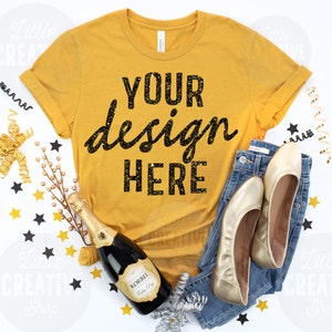May include: A mustard yellow t-shirt with the text "YOUR design HERE" in black glitter. The shirt is laying flat on a white surface with gold and black confetti, a bottle of champagne, and a pair of gold flats.