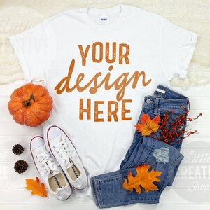 May include: White t-shirt with the text "YOUR design HERE" in orange. The shirt is laid flat on a white wooden surface with a pair of white sneakers, a small pumpkin, and a pair of blue jeans with fall leaves.