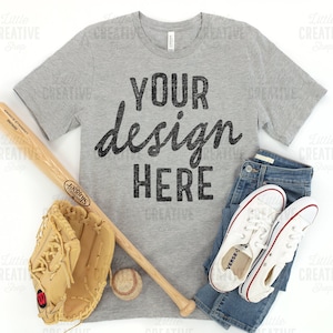 May include: A gray t-shirt with the text "YOUR design HERE" in black. The shirt is laid flat on a white surface with a baseball bat, a baseball glove, a baseball, and a pair of white sneakers and blue jeans.