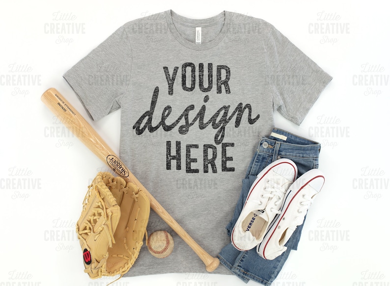 May include: A gray t-shirt with the text "YOUR design HERE" in black. The shirt is laid flat on a white surface with a baseball bat, a baseball glove, a baseball, and a pair of white sneakers and blue jeans.
