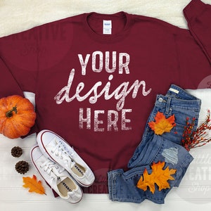 May include: A maroon sweatshirt with the text "YOUR design HERE" in white on the front. The sweatshirt is laid flat on a white surface with a pair of white sneakers, a pair of blue jeans, a small pumpkin, and fall leaves.