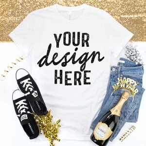 May include: White t-shirt with the text "YOUR design HERE" in black. The shirt is laying flat on a white surface with a pair of black sneakers, a bottle of champagne, a pair of blue jeans, and gold confetti.