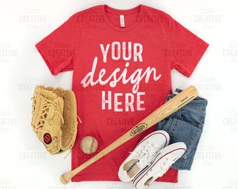 May include: Red t-shirt with the text "YOUR design HERE" in white. The shirt is laid flat on a white surface with a baseball glove, bat, and sneakers.