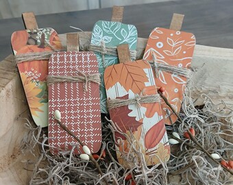 May include: A collection of decorative wooden pumpkin-shaped ornaments. Each pumpkin features a unique design, including floral and plaid patterns, and is wrapped with twine. The pumpkins are displayed in a wooden bowl with decorative moss and berries.