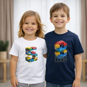 Könnte beinhalten: Zwei Kinder tragen weiße und marineblaue T-Shirts. Das weiße Shirt hat eine bunte Zahl 5 aus Spielzeugblöcken. Das marineblaue Shirt hat eine Zahl 8 aus Spielzeugblöcken und den Namen "ELIAN" in Gelb.