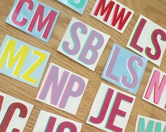 May include: A collection of colorful, square stickers with letters printed on them. The letters are in various colors, including pink, blue, red, yellow, and green. The stickers are arranged in a random pattern on a wooden surface.