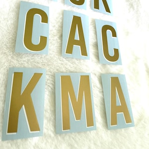May include: Gold-colored uppercase letters on light blue rectangular stickers. The letters are arranged in a scattered pattern. The stickers are on a white, fluffy surface. The letters spell out the word "ACKCACKMA".