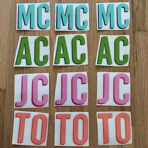 May include: Nine stickers with two-letter combinations: MC in teal and blue, AC in green, JC in pink, and TO in orange. Each sticker is on a white background, arranged in three rows of three on a wooden surface.
