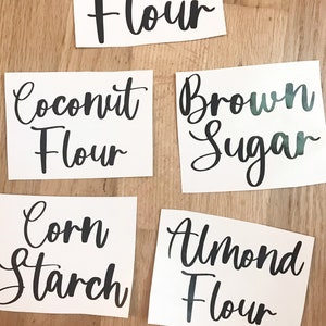 May include: White rectangular labels with black cursive text. The labels read "Flour", "Coconut Flour", "Brown Sugar", "Corn Starch", and "Almond Flour". The labels are arranged on a light brown wooden surface.