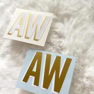 May include: Two stickers with the letters "AW" in a layered, bold font. The sticker on the left is white with gold lettering, and the sticker on the right is blue with gold lettering.
