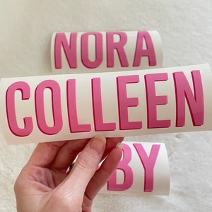 May include: Three pink vinyl stickers with the names "Nora", "Colleen", and "By" written in a bold, sans-serif font.