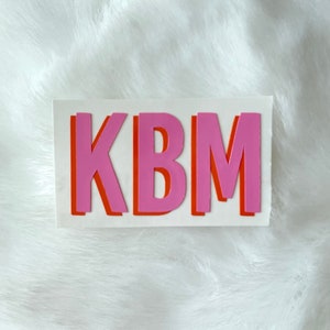 Op de afbeelding: Een witte sticker met de letters KBM in roze en oranje. De letters zijn omrand met oranje en roze, wat een gelaagd effect cre&euml;ert.