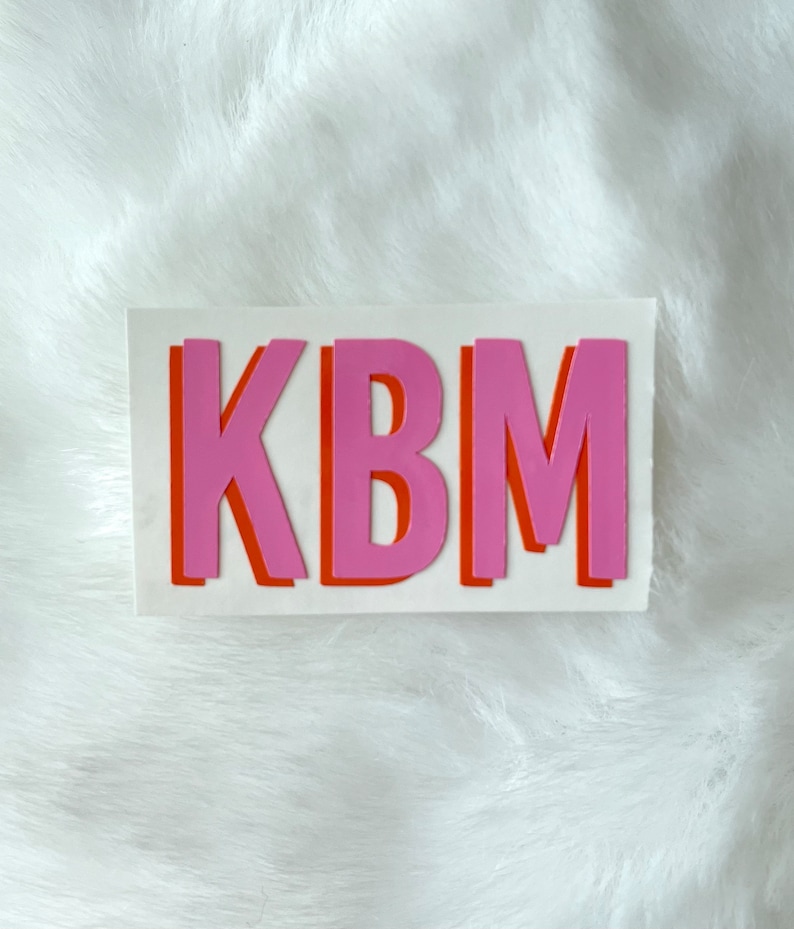 Op de afbeelding: Een witte sticker met de letters KBM in roze en oranje. De letters zijn omrand met oranje en roze, wat een gelaagd effect cre&euml;ert.