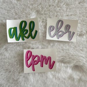 May include: Three rectangular adhesive decals featuring custom lettering in a cursive script. One decal displays the letters akl in green, one shows ebr in light purple, and one features lpm in bright pink. Each letter has a darker shadow effect.