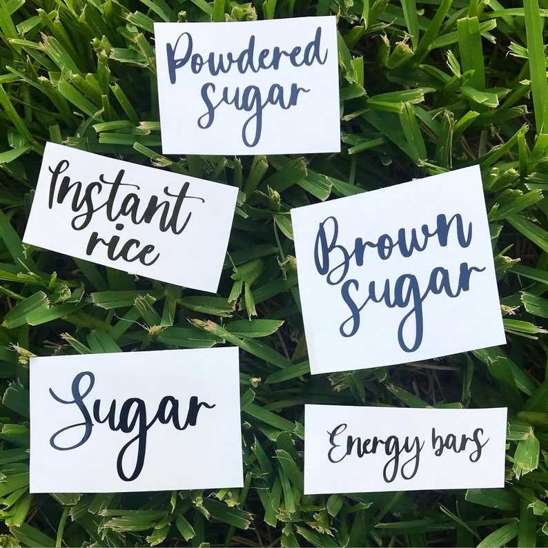 May include: Five white rectangular labels with dark blue cursive text. The labels read "Powdered Sugar", "Instant Rice", "Brown Sugar", "Sugar", and "Energy Bars". The labels are arranged on a bed of green grass.